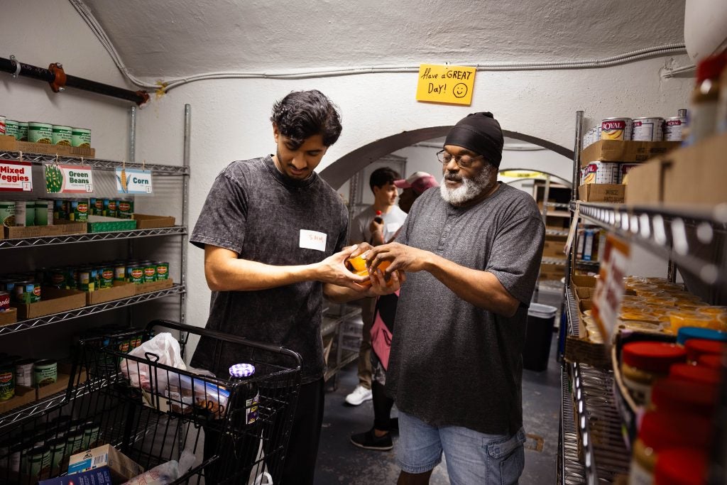 A student passes a food item to another man. They are surrounded by shelves of groceries in a food pantry