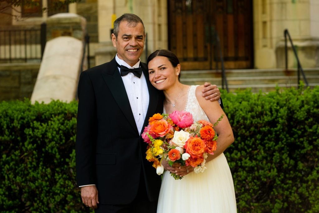 A father of the bride dressed in a tux poses with his daughter in a wedding dress holding bright orange, pink and yellow flowers
