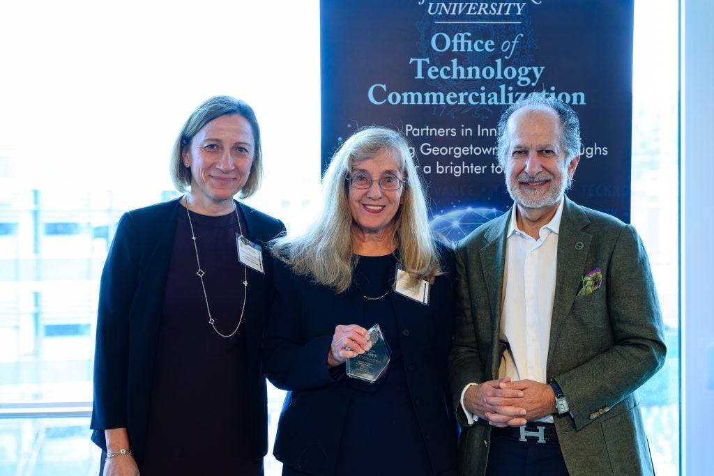 Three people pose for a picture behind a blue sign that says "Office of Technology Commercialization." The woman in the middle holds a glass trophy