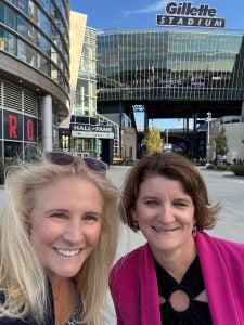 Two women smile for a selfie in front of Gillette Stadium
