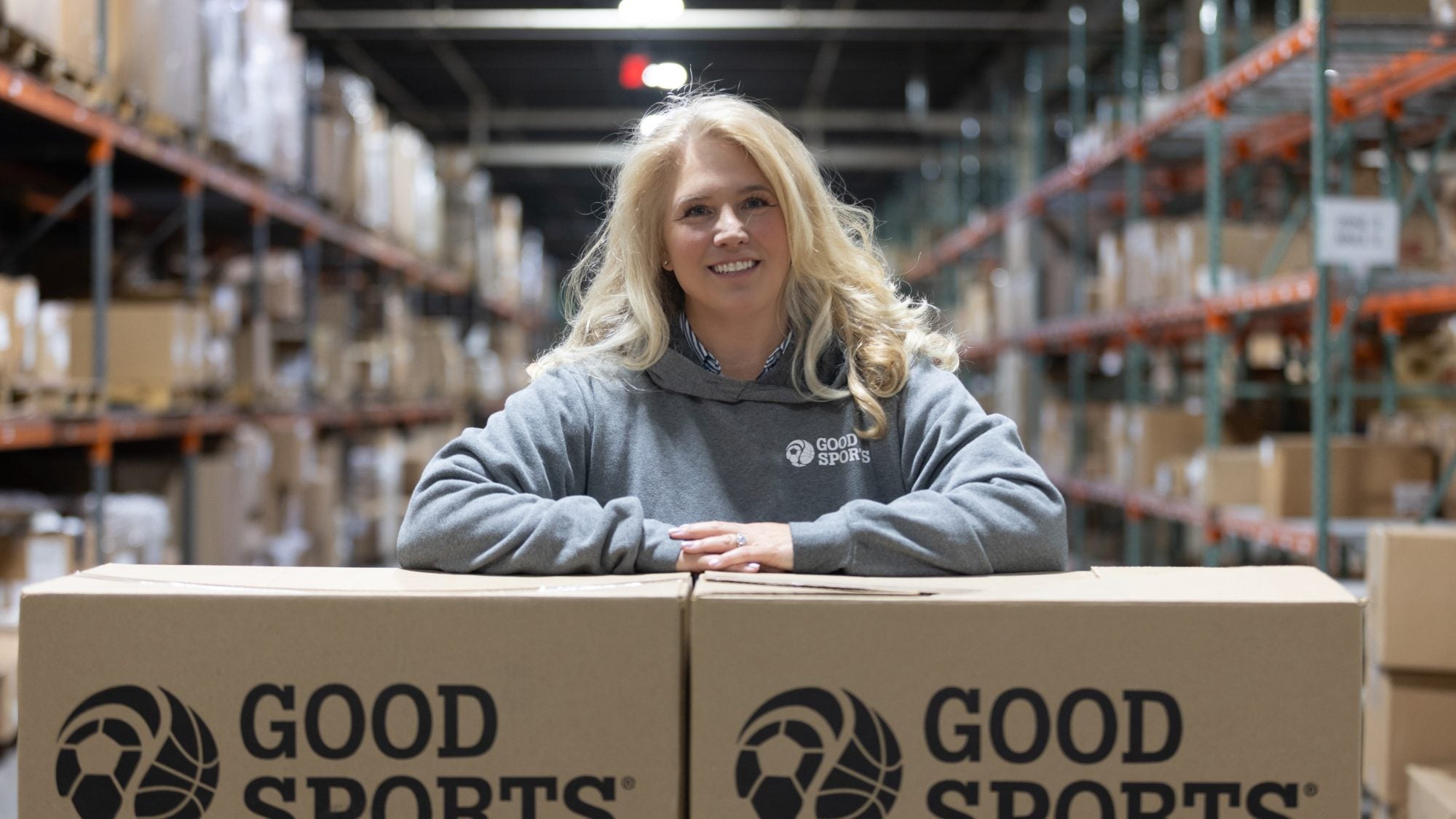 A blond woman with arms crossed over Good Sports boxes in a warehouse