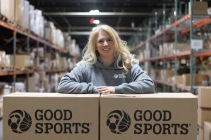 A blond woman with arms crossed over Good Sports boxes in a warehouse