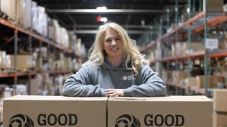 A blond woman with arms crossed over Good Sports boxes in a warehouse
