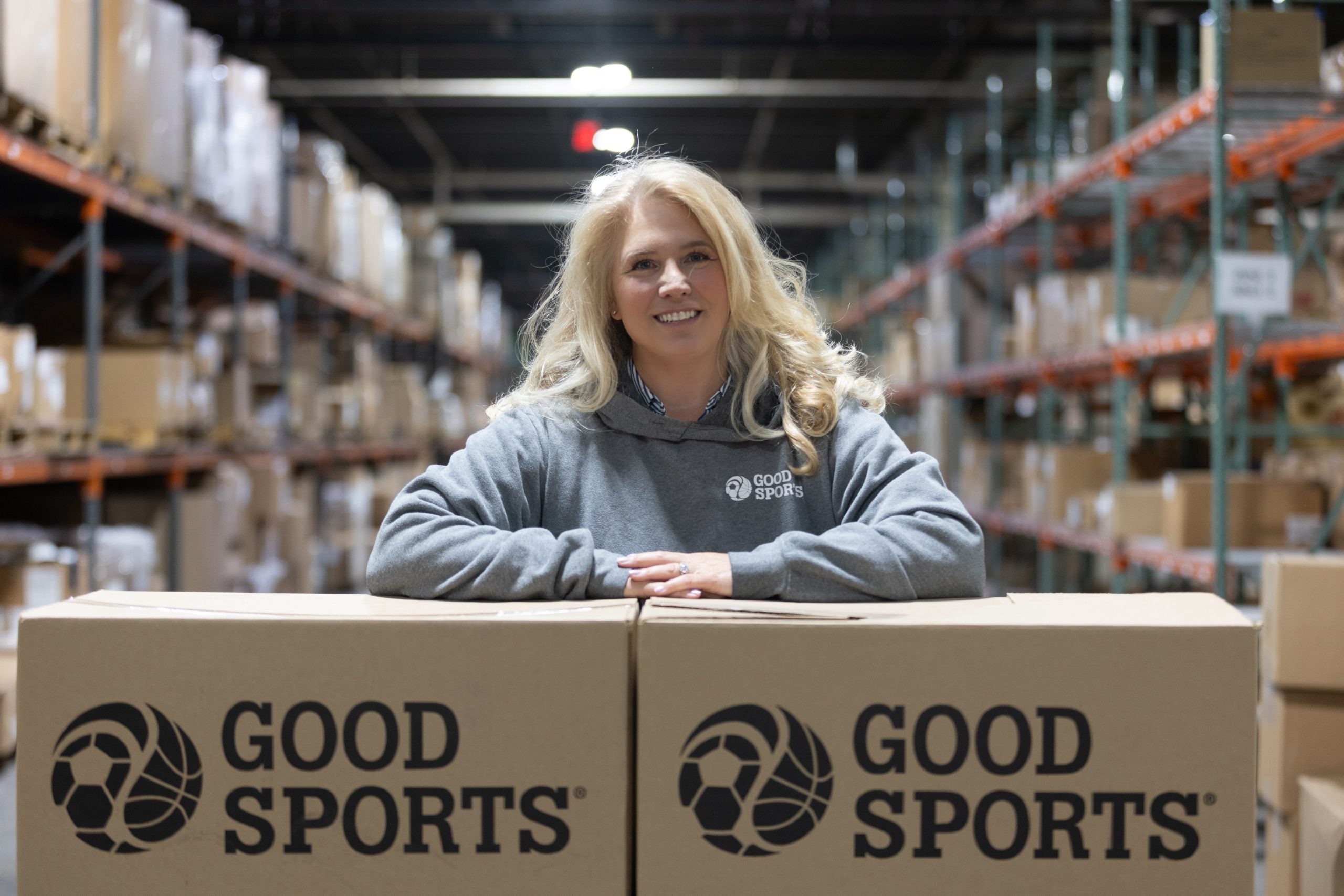 A blond woman with arms crossed over Good Sports boxes in a warehouse