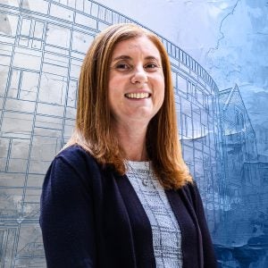 A woman with red hair in a suit smiles in front of a textured blue drawing of a building