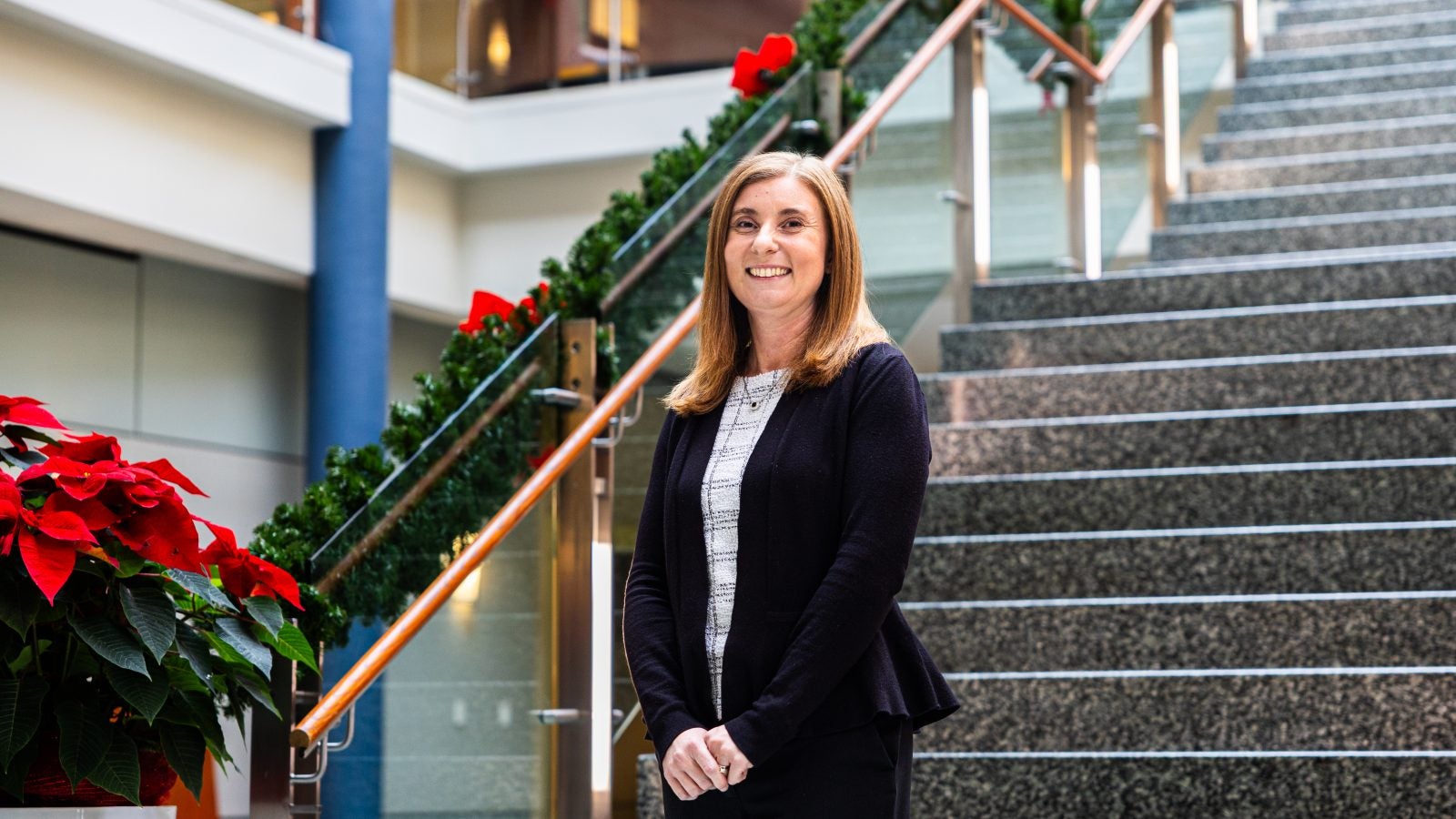 A woman in a black suit jacket and red hair smiles in front of a staircase during the holiday season