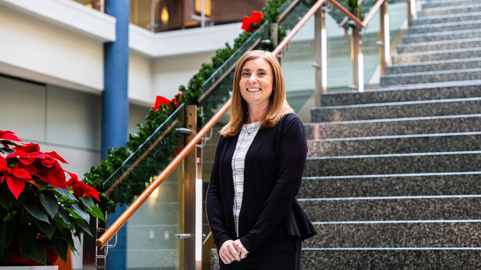 A woman in a black suit jacket and red hair smiles in front of a staircase during the holiday season