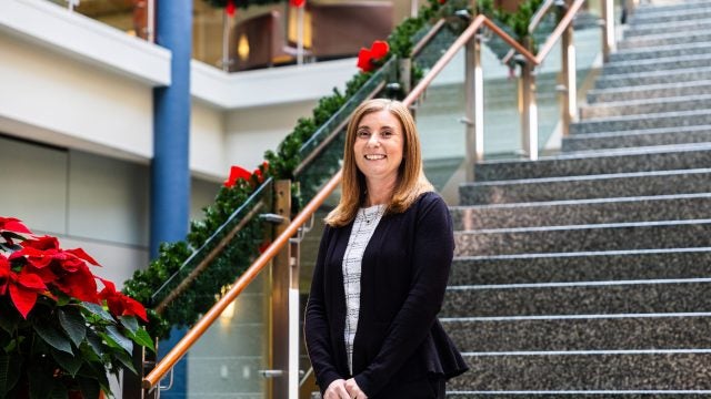 A woman in a black suit jacket and red hair smiles in front of a staircase during the holiday season