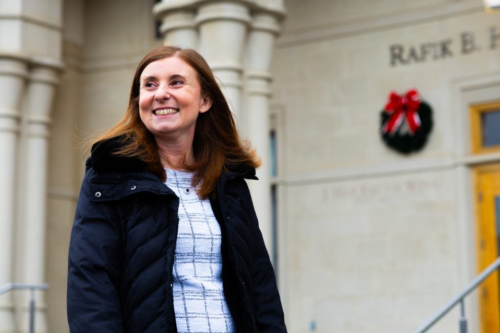 A woman in a coat smiles in front of a building with a Christmas wreath on it