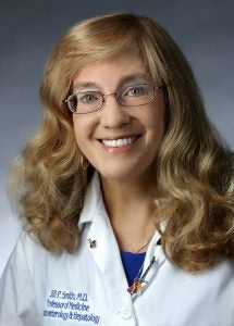 A doctor smiles in her white coat. On the collar are angel pins and a guitar