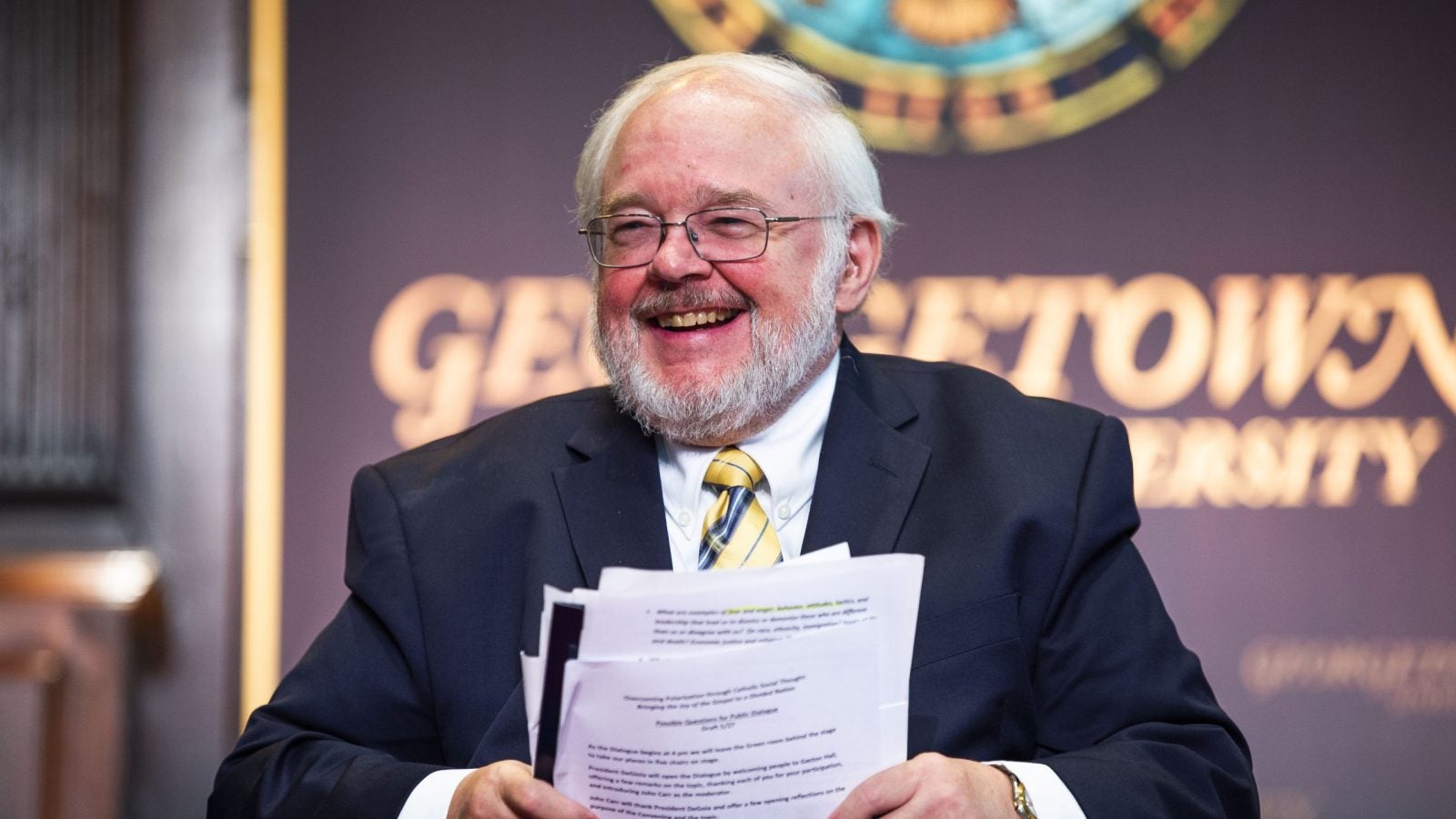 A man wearing glasses and with white hair and a beard smiles onstage while holding papers.