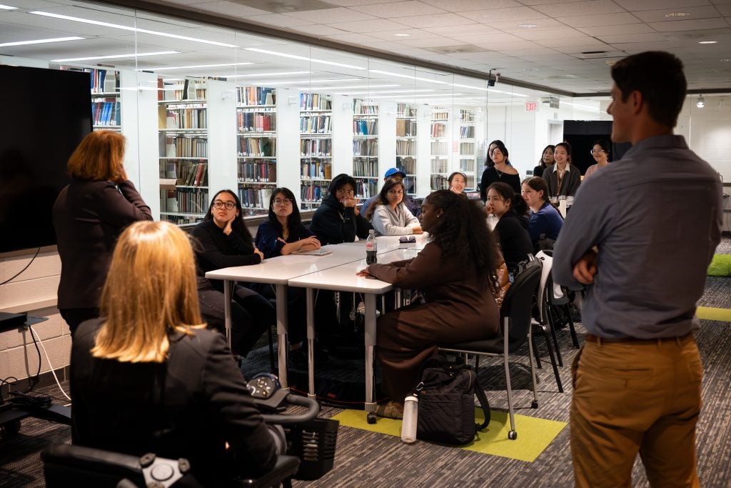 A group of students gather around a table. A woman in a mobility scooter faces them and two others stand next to her