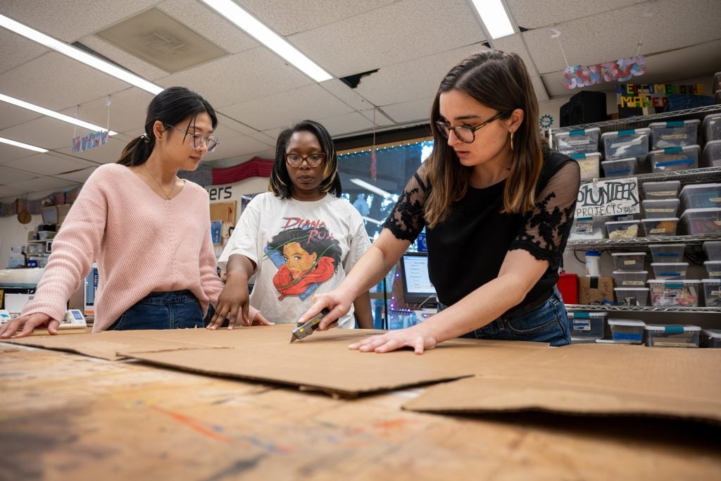 A student cuts a piece of cardboard as two other students look on