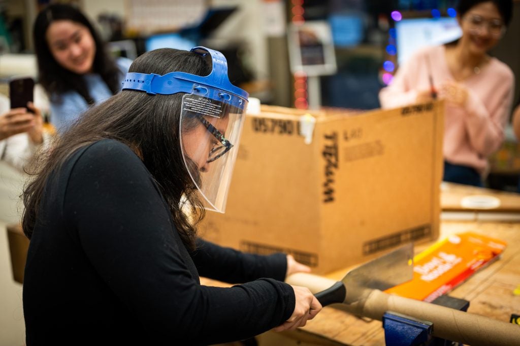A student with a plastic mask cuts into a wooden leg in a woodworking space