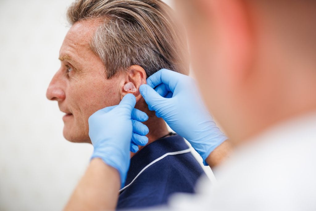 A mature Caucasian male in a blue shirt receives a personalized in-ear hearing aid from an audiologist 