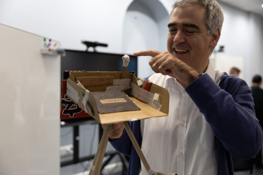 A male student smiles and points to the top of a cardboard prototype of an accessible podium, which has three legs and a mocked-up screen and microphone.