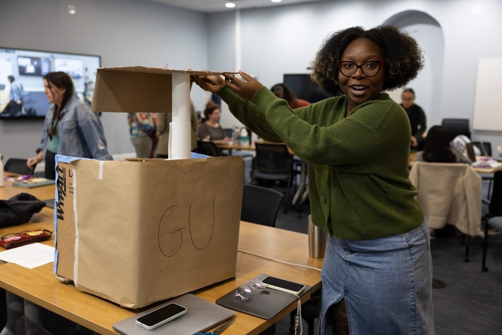 A student in a green sweater presents a cardboard prototype of an accessible podium with an adjustable base.