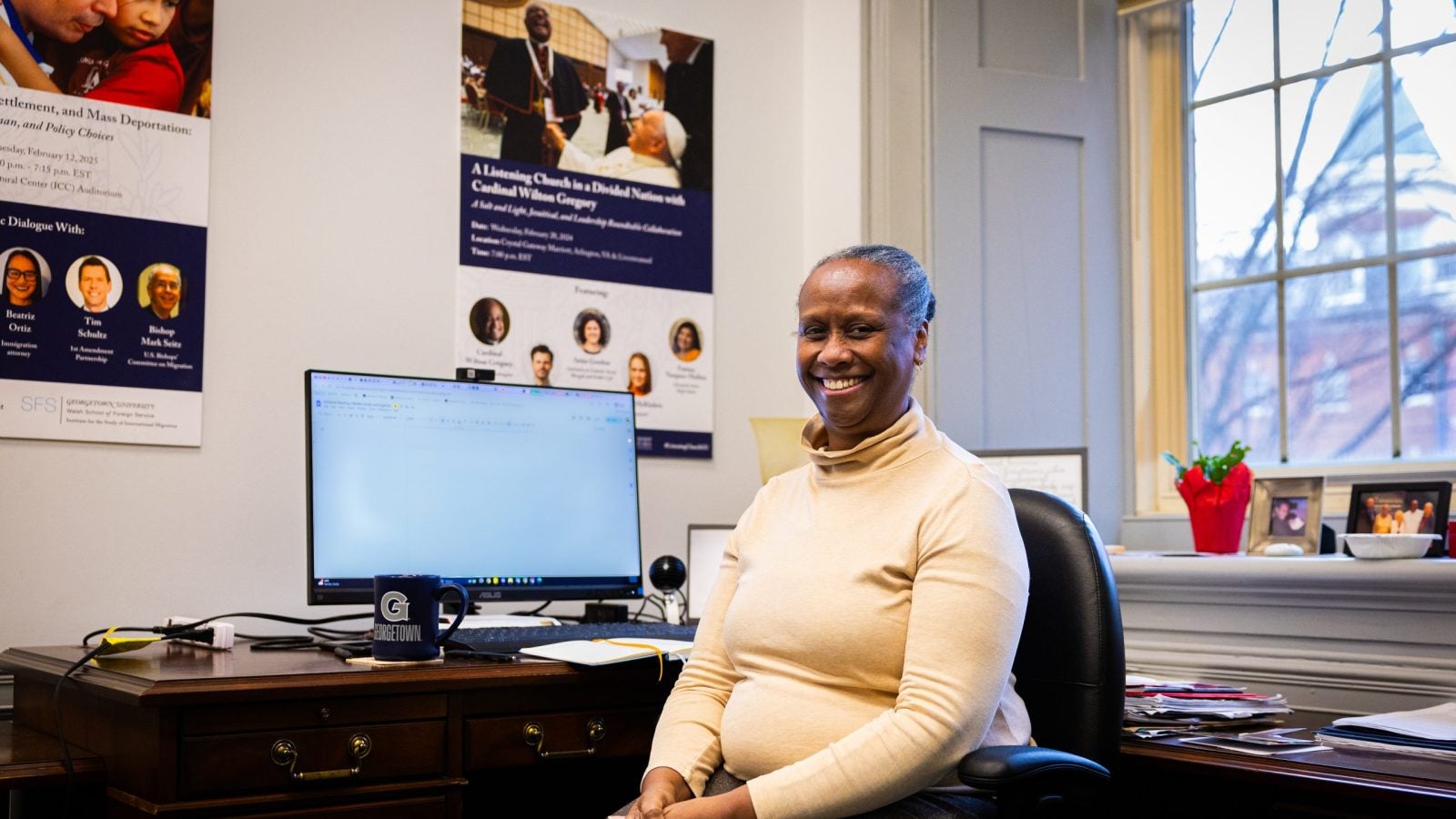 A woman in a tan turtleneck smiles at her office desk