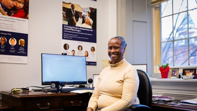A woman in a tan turtleneck smiles at her office desk