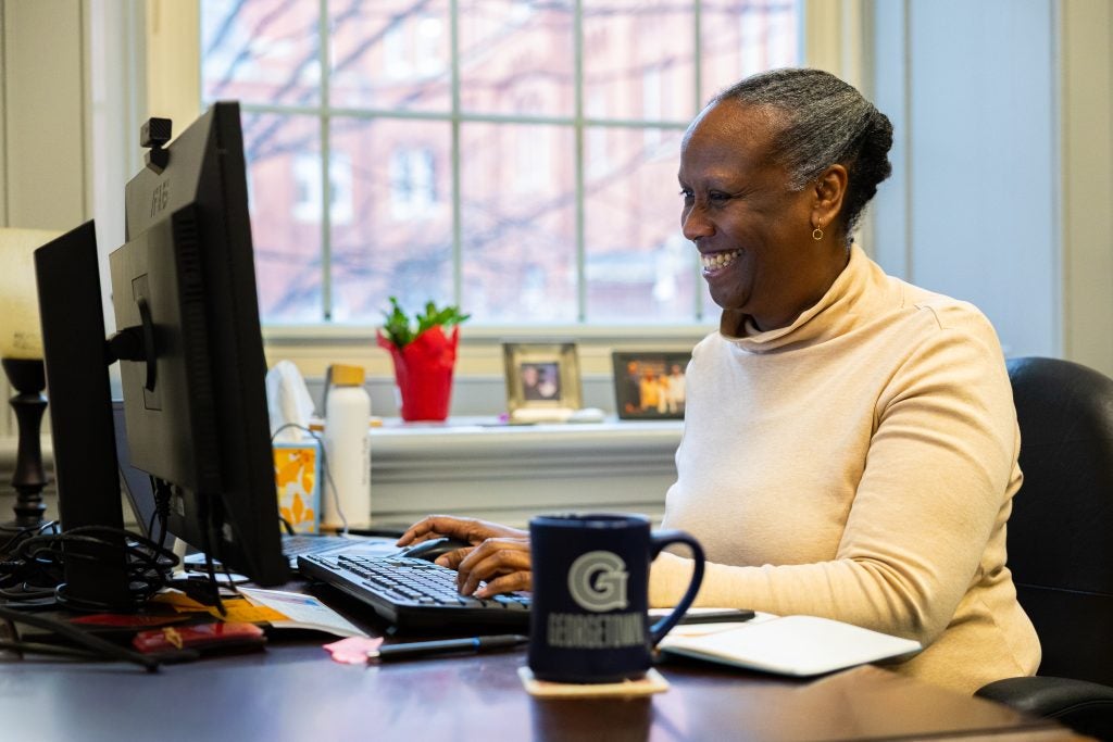 A woman smiles while working at her computer in front of a window
