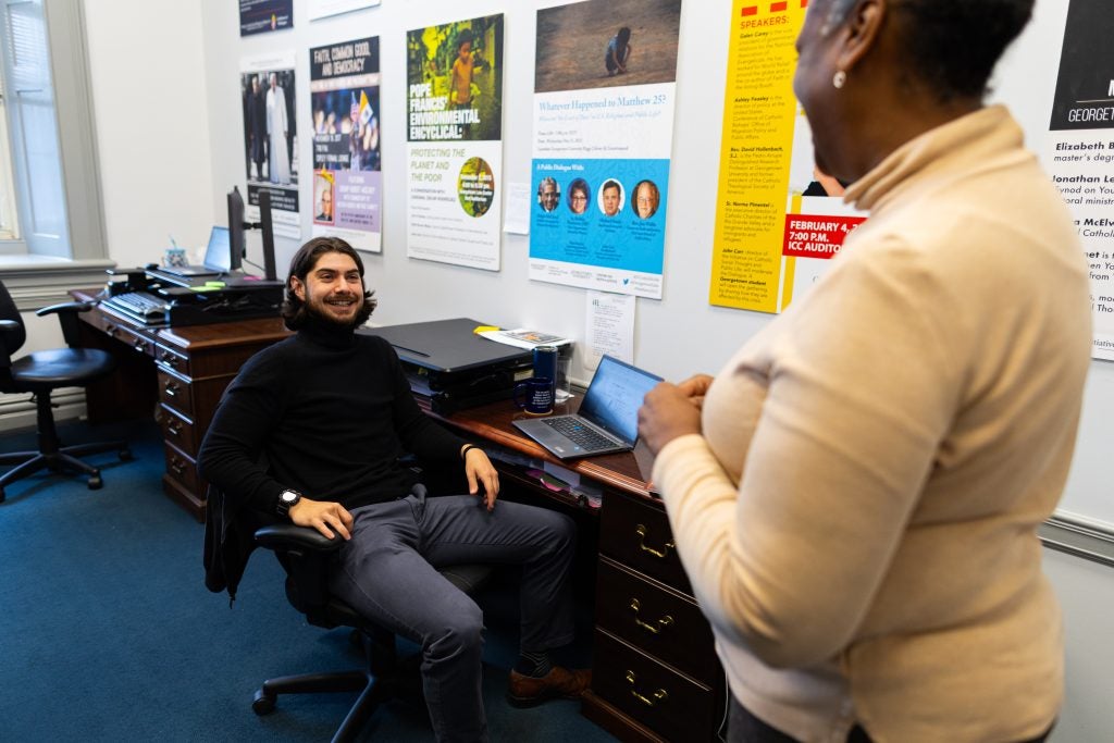 A woman speaks with her colleague, who's sitting at his office desk