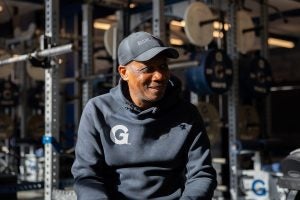 Black man smiling while wearing a Georgetown hoodie and hat and sitting down in a gym