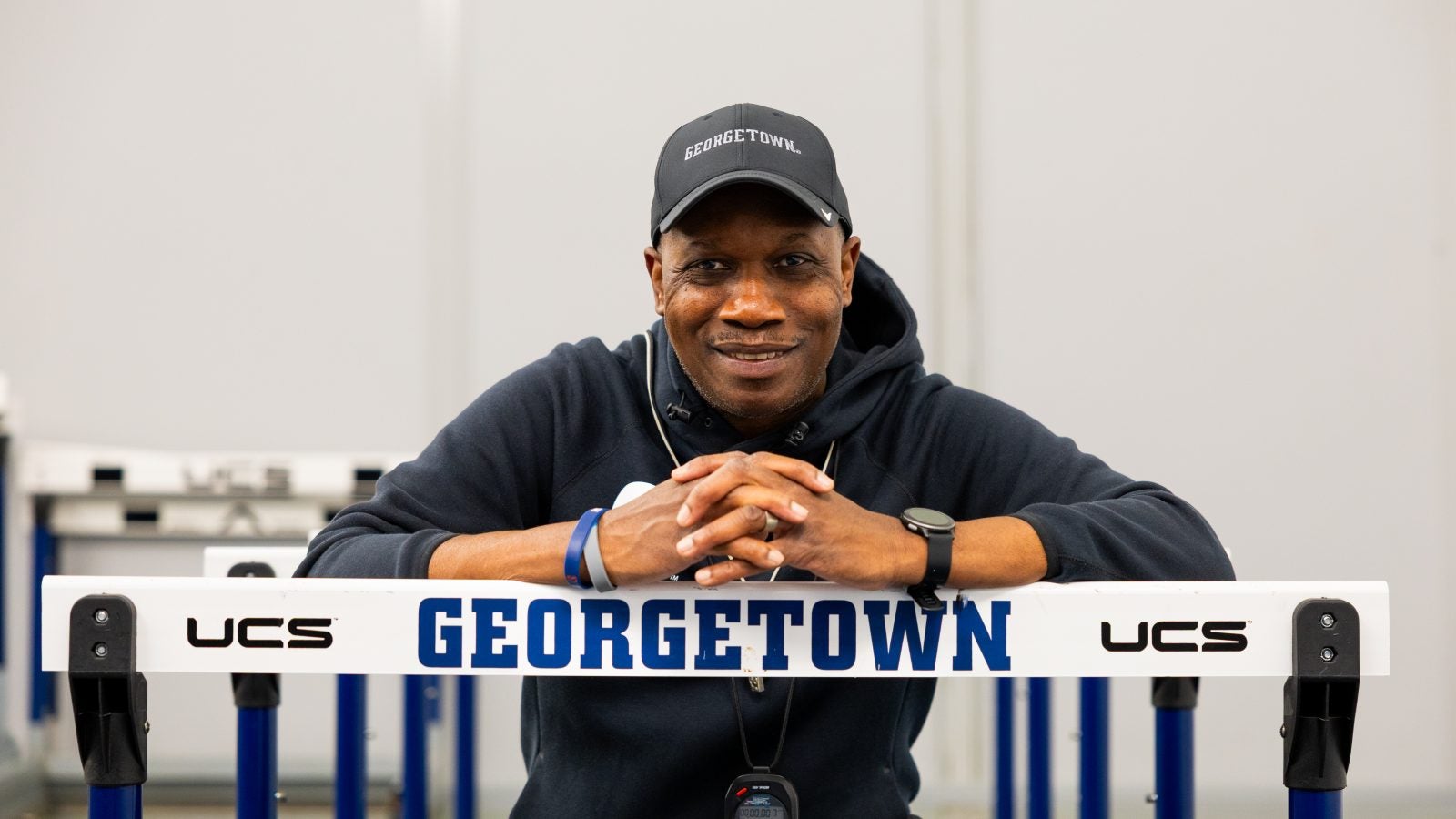 Black man smiling while wearing a Georgetown hoodie and hat and leaning over a track hurdle