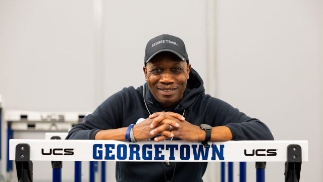 Black man smiling while wearing a Georgetown hoodie and hat and leaning over a track hurdle