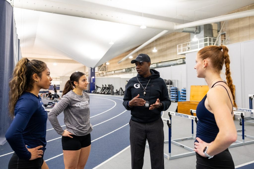 Black man wearing a Georgetown hoodie and hat coaching three young woman on track
