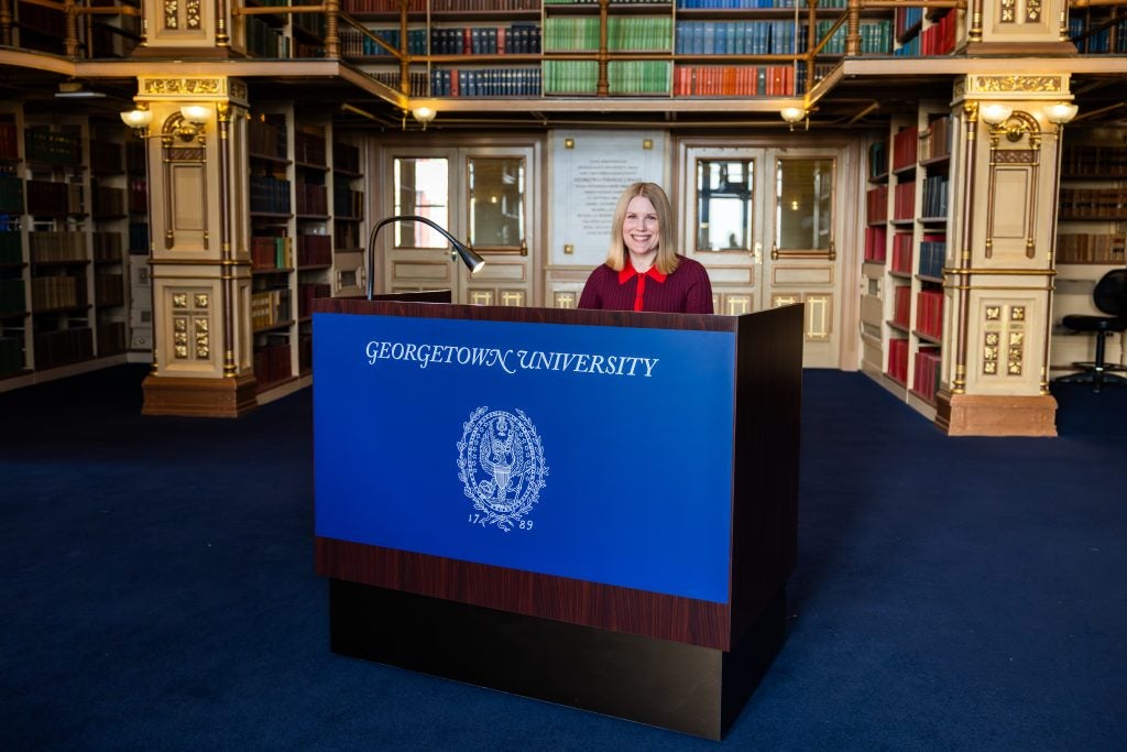 A woman sits behind an accessible brass podium with a blue sign across the front that says "Georgetown University"