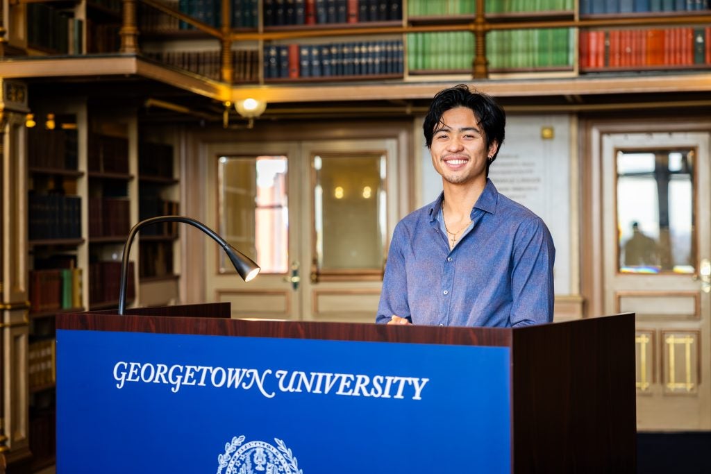 A male graduate student smiles from behind a podium that says "Georgetown University" across the front.