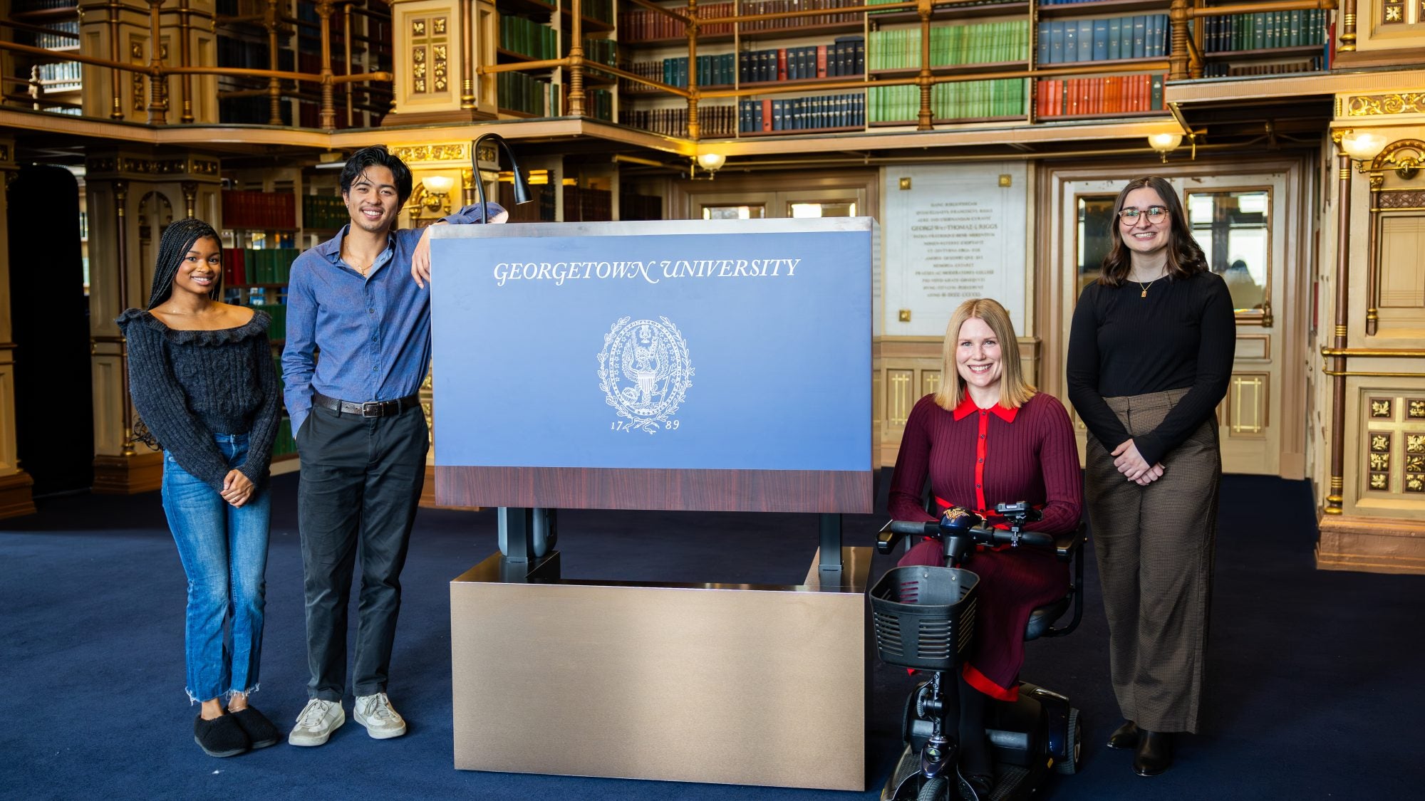 A group of students pose with an accessible podium that says "Georgetown University" across the front of it in blue. Behind them are bookshelves