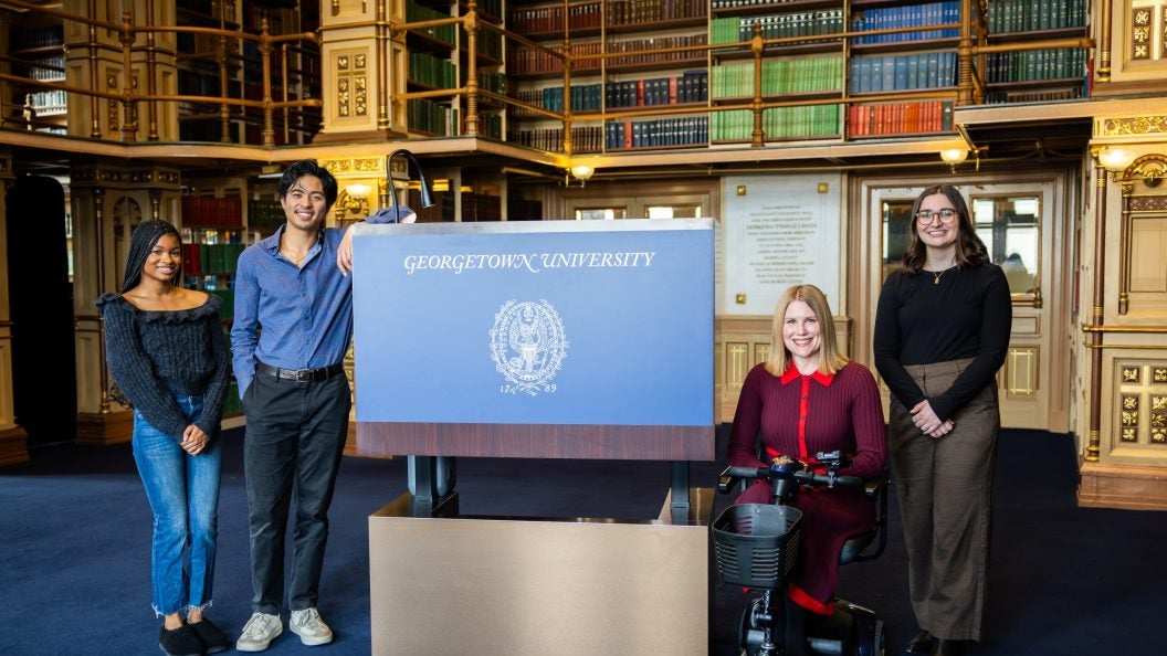 A group of students pose with an accessible podium that says "Georgetown University" across the front of it in blue. Behind them are bookshelves