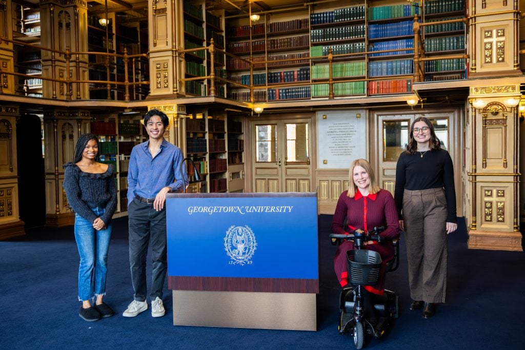 A group of students pose with an accessible podium that says "Georgetown University" across the front of it in blue. Behind them are bookshelves