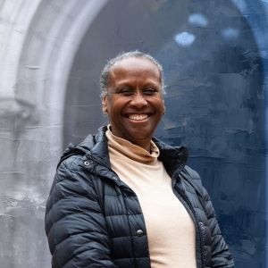 A woman smiles in front of an archway. She is wearing a black winter jacket