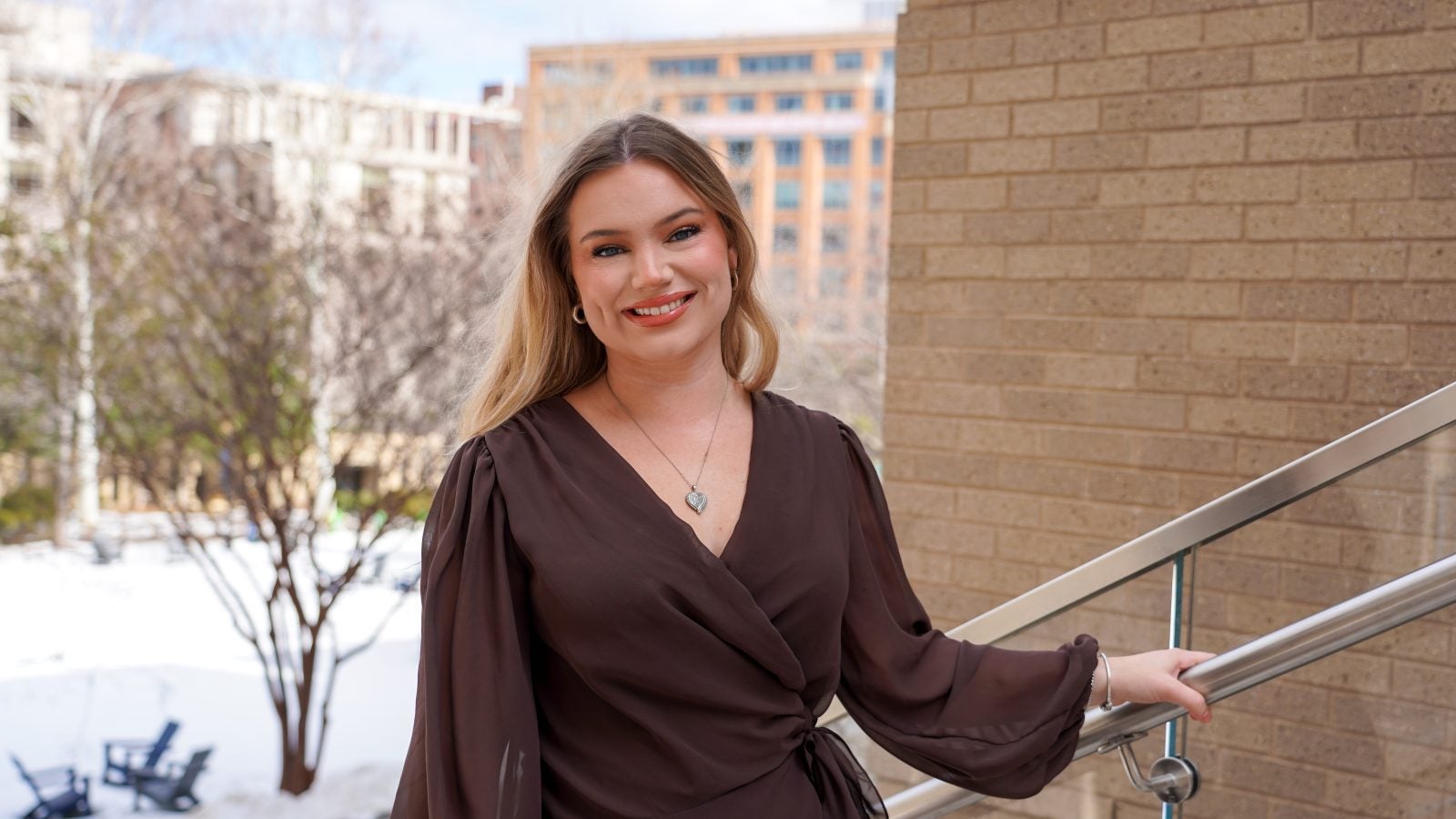 A woman in a brown shirt smiles on a stairway outdoors during the winter.