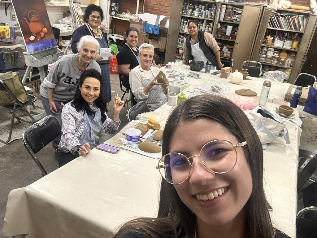 A woman in glasses takes a selfie at a ceramics studio with men and women behind her huddled around a table of pottery