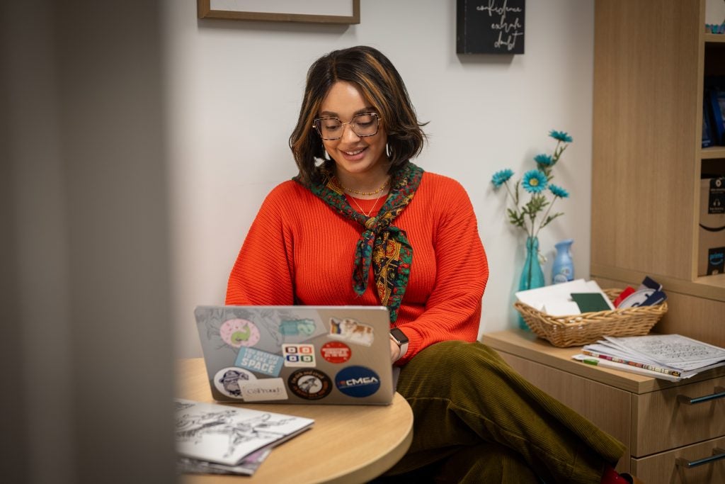 A woman in a red sweater and scarf smiles as she types on a laptop