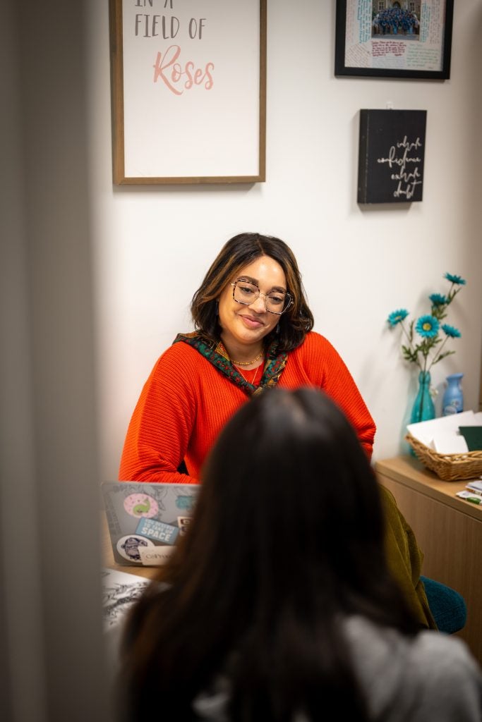 A woman in a red sweater and scarf smiles while listening to a student
