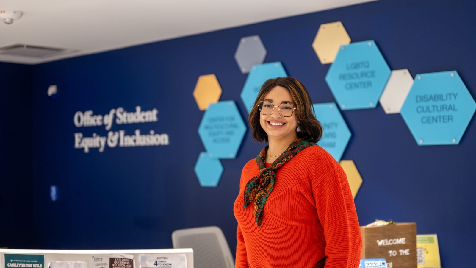A woman in a red sweater and scarf smiles in front of a navy blue wall with hexagonal shapes and a sign that says "Office of Student Equity &amp; Inclusion"