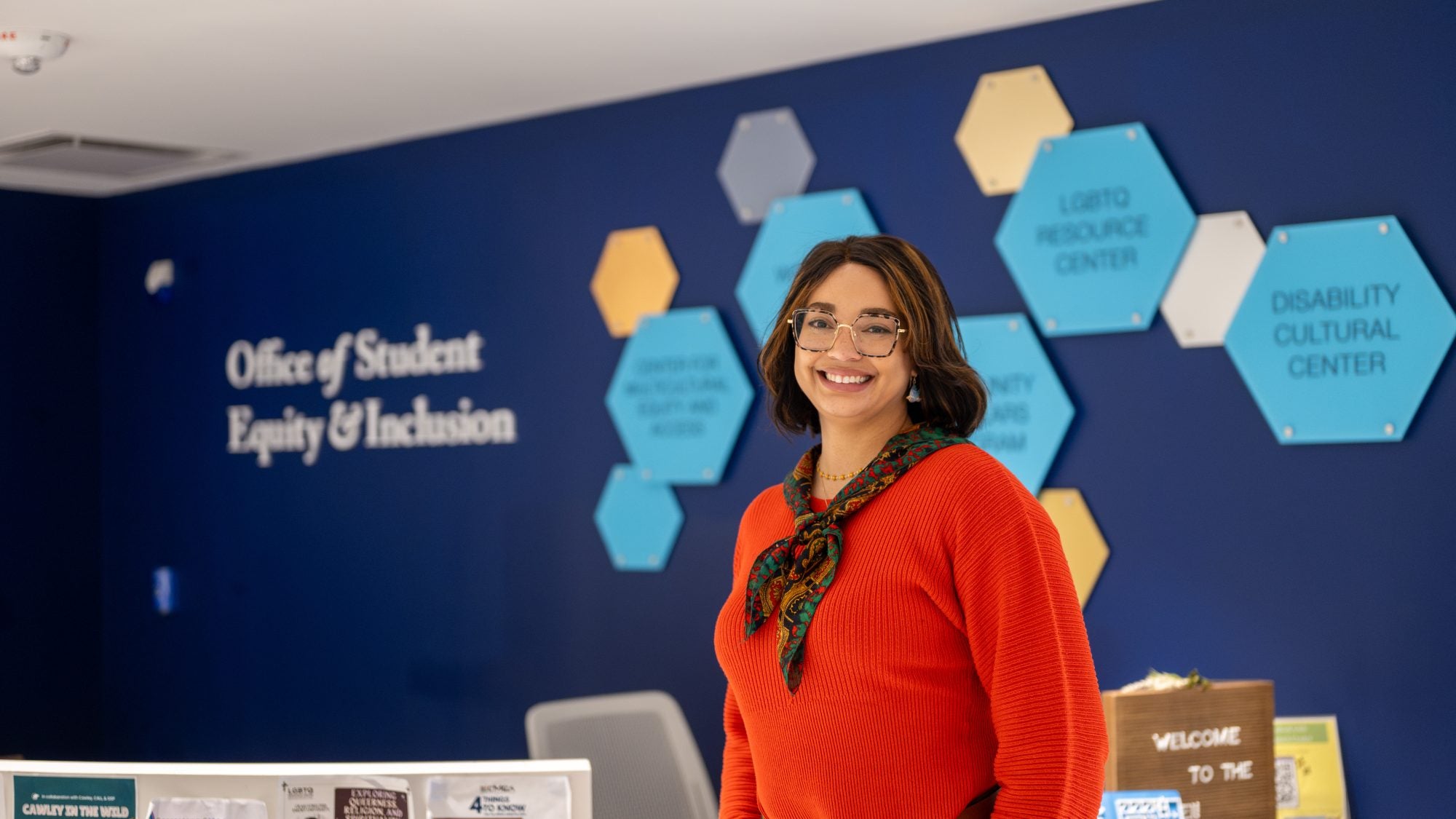 A woman in a red sweater and scarf smiles in front of a navy blue wall with hexagonal shapes and a sign that says "Office of Student Equity &amp; Inclusion"
