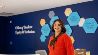 A woman in a red sweater and scarf smiles in front of a navy blue wall with hexagonal shapes and a sign that says "Office of Student Equity &amp; Inclusion"