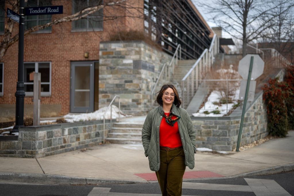 A woman in a red sweater, scarf and olive green jacket smiles while walking outside a building on a winter day