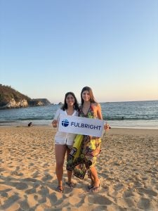 Two women stand on a beach and hold a sign that says "Fulbright"