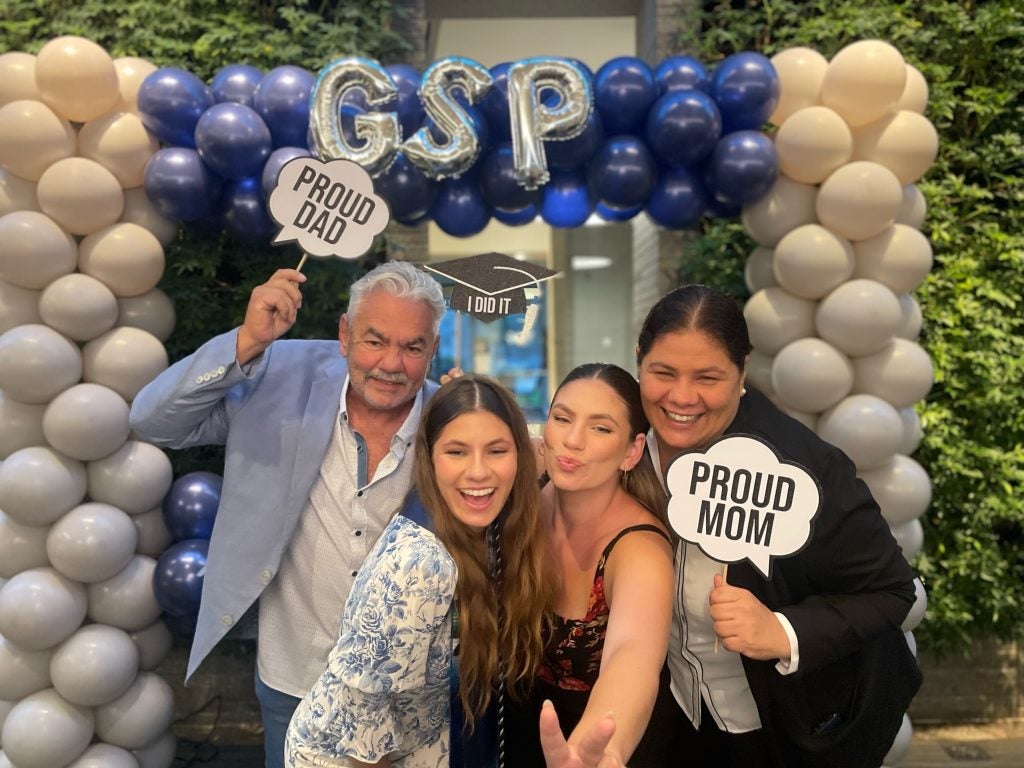 A family stands in front of balloons at a graduation ceremony. The dad and mom carry signs that say "proud dad" and "proud mom"