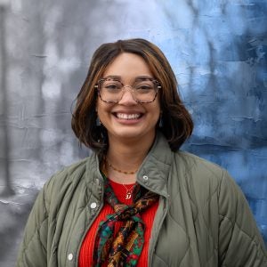 A woman in a red sweater, scarf and olive green jacket smiles in front of a blue and white background