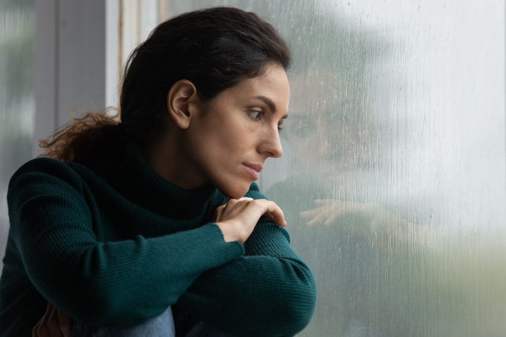 Thoughtful stressed young hispanic latin woman sitting on windowsill, looking outside on rainy weather, having depressive or melancholic mood, suffering from negative thoughts alone at home.