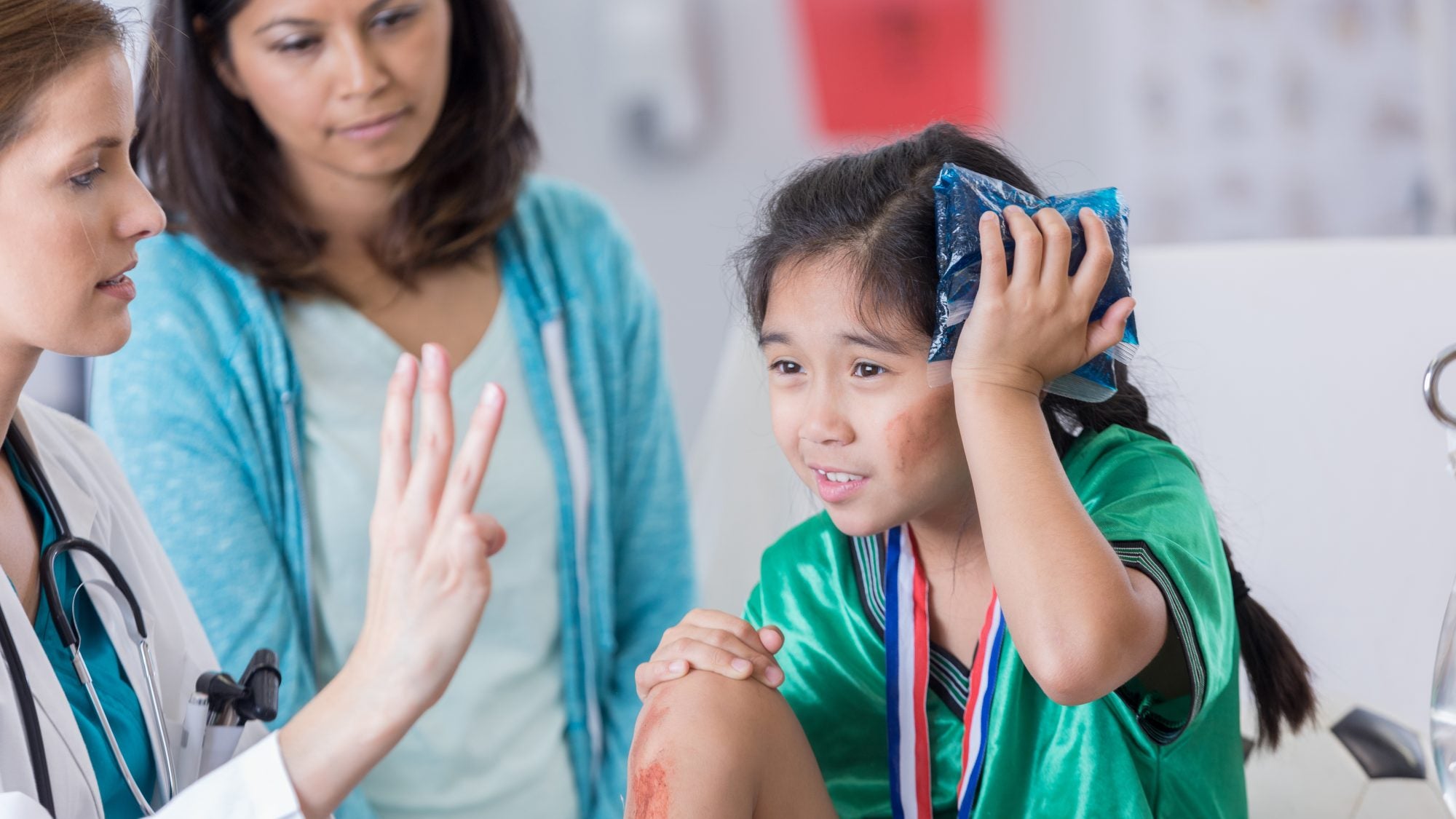 Serious female ER doctor holds up three fingers while talking with injured female elementary age soccer player. The doctor is asking the girl how many fingers she is holding up. The girl is wearing a medal and soccer uniform.
