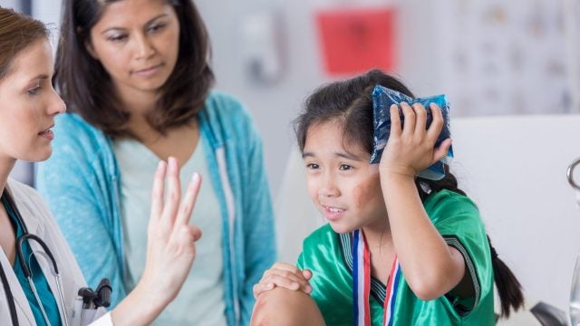 Serious female ER doctor holds up three fingers while talking with injured female elementary age soccer player. The doctor is asking the girl how many fingers she is holding up. The girl is wearing a medal and soccer uniform.