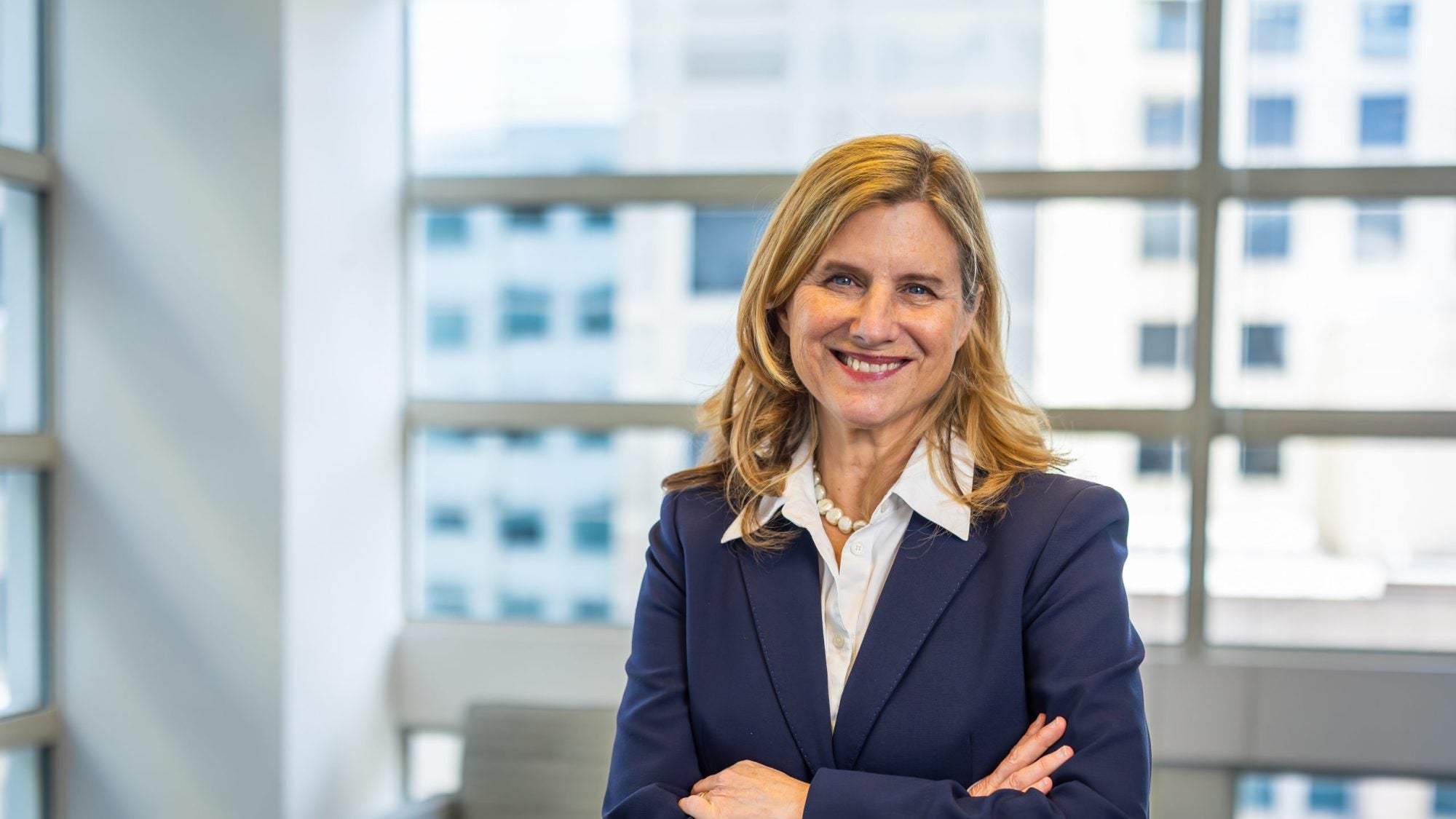 A woman in a blue suit and white shirt crosses her arms and smiles in front of windows